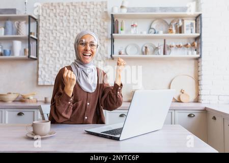 Junge, glückliche, muslimische Studentin im Hidschab, die zu Hause sitzt und auf einem Laptop lernt. Zeigt eine Ja-Geste mit den Händen an der Kamera. Ich habe eine Online-Prüfung bestanden, einen Brief von der Universität, gute Ergebnisse. Stockfoto