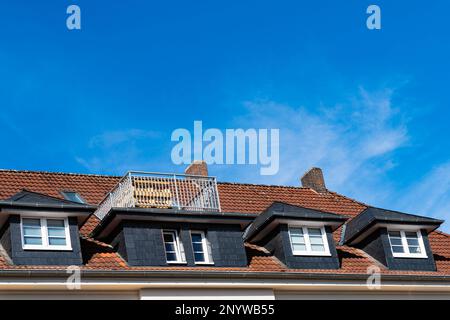 Bruchteil eines gefliesten Dachs mit Oberlichtern und einem kleinen Balkon. Blauer Himmel mit hellen Wolken. Stockfoto