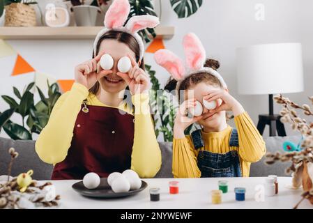 Lustige lächelnde Mutter und fröhliche, verspielte kleine Tochter, die weiße Ostereier vor die Augen hielt, während sie sie im Wohnzimmer mit Lebensmittelfarbstoffen dekorierte Stockfoto