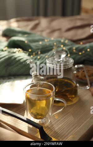 Holztablett mit frisch gebrühtem Tee auf dem Bett im Zimmer. Gemütliche Atmosphäre Stockfoto