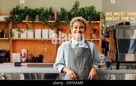 Selbstbewusster Besitzer eines Coffee Shops in einer Schürze. Junge lächelnde Frau in einer Schürze, die an der Bar steht. Stockfoto