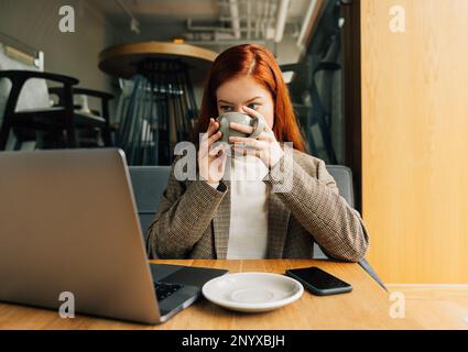 Frau trinkt Kaffee und schaut auf Laptop, während sie an einem Tisch im Café sitzt Stockfoto