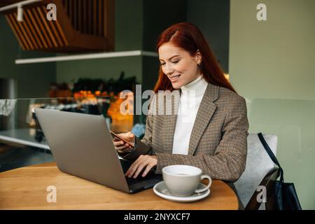 Lächelnde rothaarige Frau, die auf einem Laptop tippt und ein Handy hält, während sie in einem Café sitzt Stockfoto
