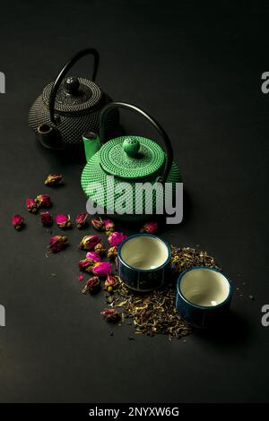 Ein Stillleben mit zwei zweifarbigen gusseisernen Teekannen zusammen mit ein paar kleinen Porzellanbechern und getrocknetem Rosentee. Stockfoto