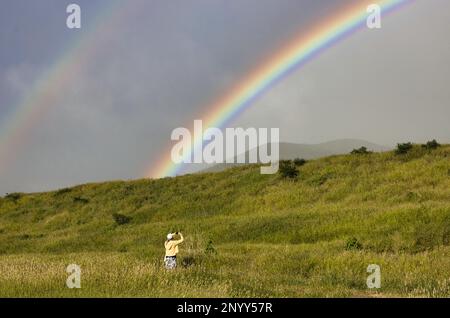 Unidentifizierbare Frau, die ein Handyfoto von einem Regenbogen über den Bergen von West maui aufnimmt. Stockfoto