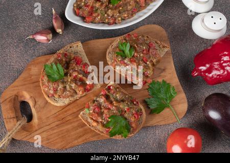 Hausgemachter Auberginen-Kaviar mit Pfeffer, Tomaten und Knoblauch auf Toast auf Holzbrett, Blick von oben Stockfoto
