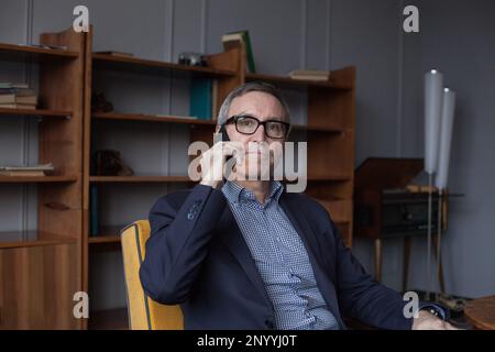 Porträt eines älteren, selbstbewussten, grauhaarigen Geschäftsmanns in blauem Anzug und Brille, der mit dem Handy auf grauem Studiohintergrund spricht Stockfoto
