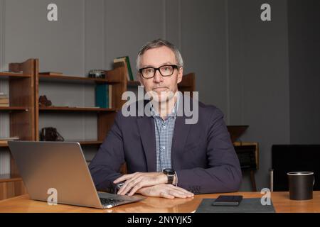 Ein erwachsener, grauhaariger Geschäftsmann oder Unternehmer in Brille und Anzug sitzt neben dem Laptop in seinem Büro Stockfoto