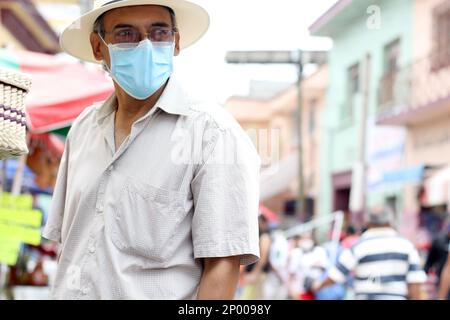 Lateinamerikanischer älterer Erwachsener mit Gesichtsschutzmasken und Hut beim Gehen in plaza, neuer normaler Covid-19 Stockfoto