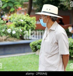 Lateinamerikanischer älterer Erwachsener mit Gesichtsschutzmasken und Hut beim Gehen in plaza, neuer normaler Covid-19 Stockfoto