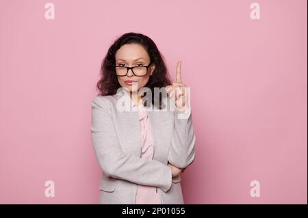 Eine selbstbewusste, ethnische Frau, die eine Brille und einen formellen Anzug trägt, zeigt mit dem Zeigefinger nach oben auf die Textstelle auf pinkfarbenem Hintergrund Stockfoto