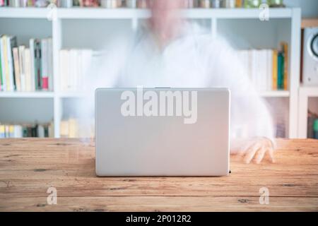 Verschwommener erwachsener Mann, der von zu Hause aus vor einem Laptop an einem Holztisch und einem Bücherregal voller Bücher in einem hellen Raum arbeitet Stockfoto