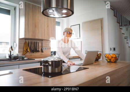 Blonde weiße Frau, die in der Küche an ihrem Laptop arbeitet Stockfoto