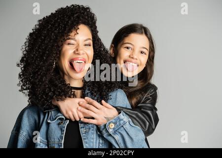 Lockige Frau und Tochter, die aus den Zungen herausragen, isoliert auf grauem, Stock-Bild Stockfoto