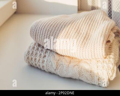 Haufen Strickpullover mit Zopfmuster auf sonniger Fensterbank. Zwei warme Strickjacken. Kuscheltes Kleidungsstück für die kalte Jahreszeit. Stockfoto