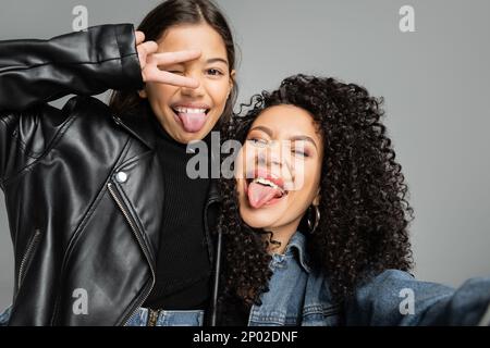 Trendige Frau und Tochter, die ihre Zungen ausrasten, isoliert auf grauem Aktienbild Stockfoto