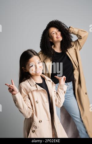 Lächelndes Mädchen im Trenchcoat zeigt Friedenszeichen in der Nähe von mom isoliert auf grauem Stockbild Stockfoto