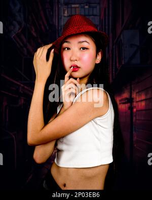 Teenager-Mädchen mit roter Fedora, die in der Gasse steht, mit Händen auf den Lippen und Hut | Rote Töne | Moody Stockfoto