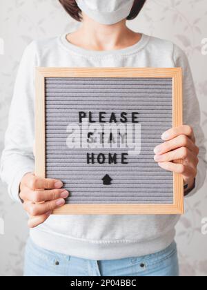 Frau in medizinischer Maske mit Briefbrett. SATZ, BITTE ZU HAUSE BLEIBEN. Quarantäne wegen Coronavirus COVID19. Selbstisolierung zu Hause. Stockfoto