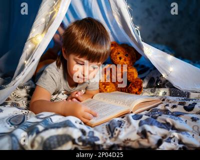 Der kleine Junge liest mit seinem Teddybär ein Buch. Kleinkind spielt im Zelt aus Leinenlaken auf dem Bett. Gemütlicher Abend mit Lieblingsbuch. Stockfoto