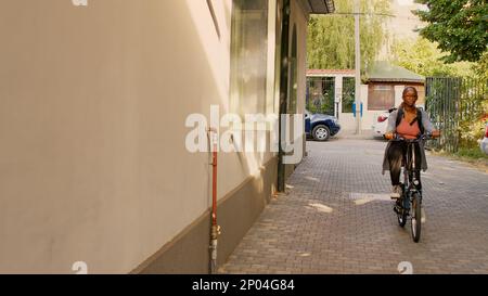 Eine junge Kurierin auf dem Fahrrad, die zum Kundeneingang geht, um den Restaurantbestellservice zu liefern. Junge Lieferperson, die Fahrrad fährt und Kunden Fastfood gibt, Tragetasche. Stockfoto