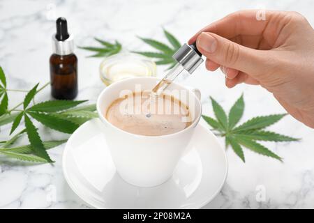 Frau tropft THC-Tinktur oder CBD-Öl in eine Tasse Kaffee am weißen Marmortisch, Nahaufnahme Stockfoto