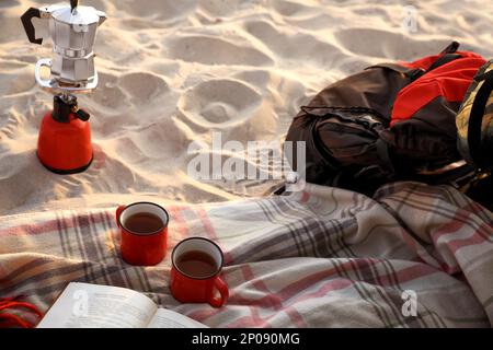 Tassen Tee, buchen Sie auf einer Decke in der Nähe des Campingkochers mit Moka-Topf und Rucksack am Sandstrand Stockfoto