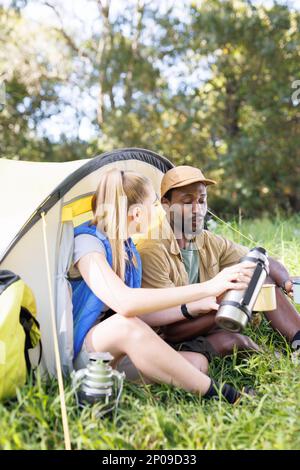 Ein glückliches, vielseitiges Paar im Wald, das vor dem Zelt sitzt, Kaffee eingießt, Kopierraum Stockfoto