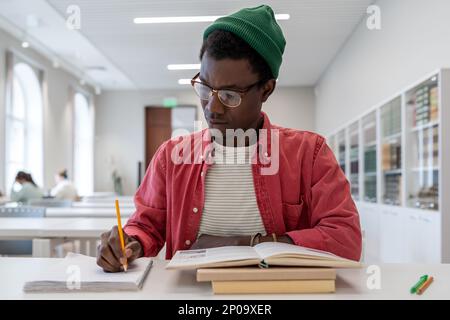 Fokussierter afroamerikanischer Student, der am Schreibtisch sitzt, mit Lehrbüchern, in der Bibliothek lernt Stockfoto
