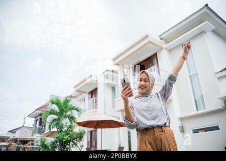 Aufgeregte Frau im Hijab mit Handy im häuslichen Umfeld Stockfoto