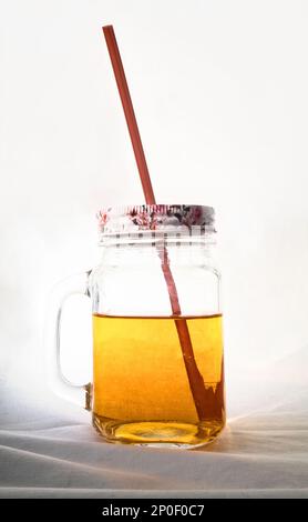 Klar Einmachglas Apfelsaft mit einem Strohhalm und einem Deckel isoliert auf weiss. Gelbe Getränk in einem transparenten Glas-Becher Stockfoto