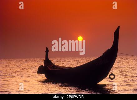 Die untergehende Sonne umrahmt von dem sichelförmigen Heck eines Katamarans in Kozhikode, Calicut, Kerala, Indien Stockfoto