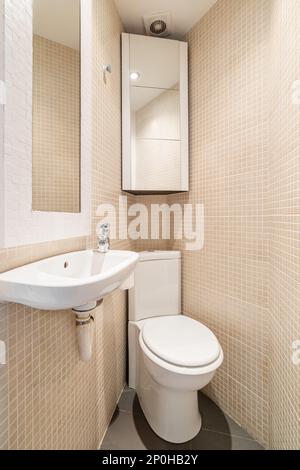 Blick auf die Toilette in der Ecke des kompakten, konischen Badezimmers mit beigefarbenen Mosaikfliesen, Spiegel, Toilette, Waschbecken und Wandschrank. Design-Konzept Stockfoto