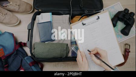 Nahaufnahme der Reise-Checkliste und verpackter Koffer und weibliche Handprüfungsgegenstände, die sich für die Reise vorbereiten. Tourismus- und Gepäckvorbereitungskonzept. Stockfoto