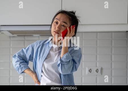Verärgerte, unzufriedene junge Asiatin, die zu Hause telefoniert und sich mit dem Handy streitet Stockfoto