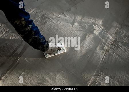 Gipsschweben in der männlichen Hand mit leuchtendem Handschuh an einer grauen Wand. Putzreibe in den Händen eines Arbeiters. Stockfoto