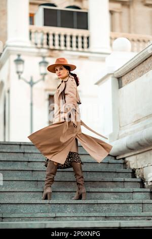 Outdoor-Modeporträt einer jungen eleganten, modischen braunen Frau, Model mit stilvollem Hut, Würfelchen und leichter Regenjacke, die bei Sonnenuntergang in Europa posiert Stockfoto