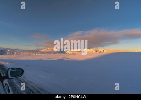 Schneebedeckte Berge, die von der Sonnenaufgangssonne Islands von der Innenansicht eines Autos beleuchtet werden, wobei ein halbes Gesicht im Rückspiegel reflektiert wird Stockfoto