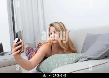 Junge blonde Frau, die Fotos mit ihrem Smartphone auf dem Sofa in ihrem Wohnzimmer macht Stockfoto