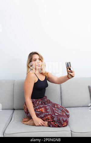 Hübsche blonde Frau, die Fotos mit ihrem Smartphone auf dem Sofa in ihrem Wohnzimmer macht Stockfoto