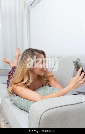 Junge blonde Frau, die Fotos mit ihrem Smartphone auf dem Sofa in ihrem Wohnzimmer macht Stockfoto