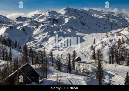 Bohinj, Slowenien - Winterblick auf den verschneiten Berg Vogel mit Skifahrern auf Skipisten und hölzernen Hütten in den Alpen im Triglav-Nationalpark auf einem sonnigen w Stockfoto