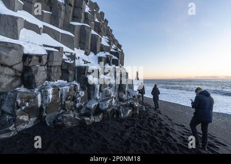 Drei Touristen spazieren entlang des Ufers des schwarzen Sandstrands Reynisfjara Beach mit den Basaltformationen zur Seite bei Sonnenuntergang beleuchtet vom Letzten Stockfoto