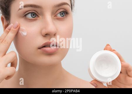 Ein hübsches Mädchen trägt Creme auf ihr Gesicht auf, Nahaufnahme. Langes Haar, große blaue Augen und klare Haut. Stockfoto