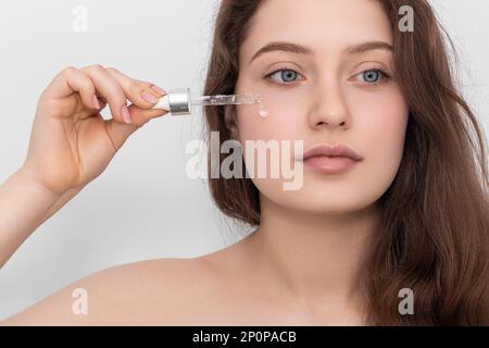 Junge Frau, die ihr Serum auf das Gesicht aufträgt und wegschaut. Stockfoto
