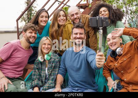 Eine Gruppe verschiedener Schüler macht ein Selfie auf einer Terrasse mit einem Selfie-Stick, einschließlich einer nicht binären Person mit rasiertem Kopf. Es gibt acht Freunde Stockfoto