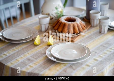 Osterzitronenkuchen, Babka mit Puderzucker auf einem festlichen Tisch, dekoriert mit Keramikschüsseln. Rustikaler, skandinavischer Stil mit Tischset Stockfoto