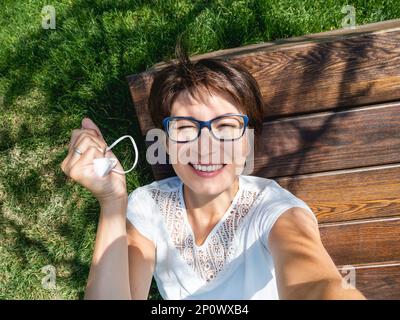 Lächelnde Frau nimmt medizinische Schutzmaske ab, um Selfie auf ihrem Smartphone zu machen. Eine Frau mit Brille hat eine Pause im Stadtpark. Neue Normalität. Stockfoto