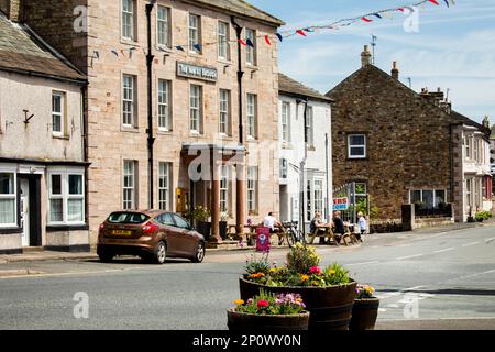 An einem sonnigen Tag in der Hauptstraße des Dorfes Brough, Cumbria, sitzen die Leute vor einem Gasthaus oder einem öffentlichen Haus, um zu essen und zu trinken Stockfoto