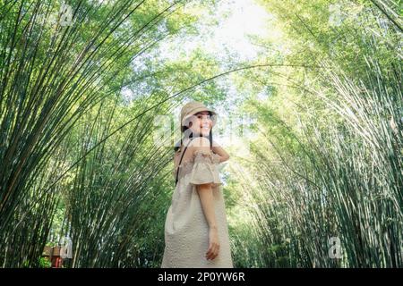 Fröhliche junge asiatische Frau mit Hut, die am helllichten Tag im Bambuswald auf dem Land stand Stockfoto